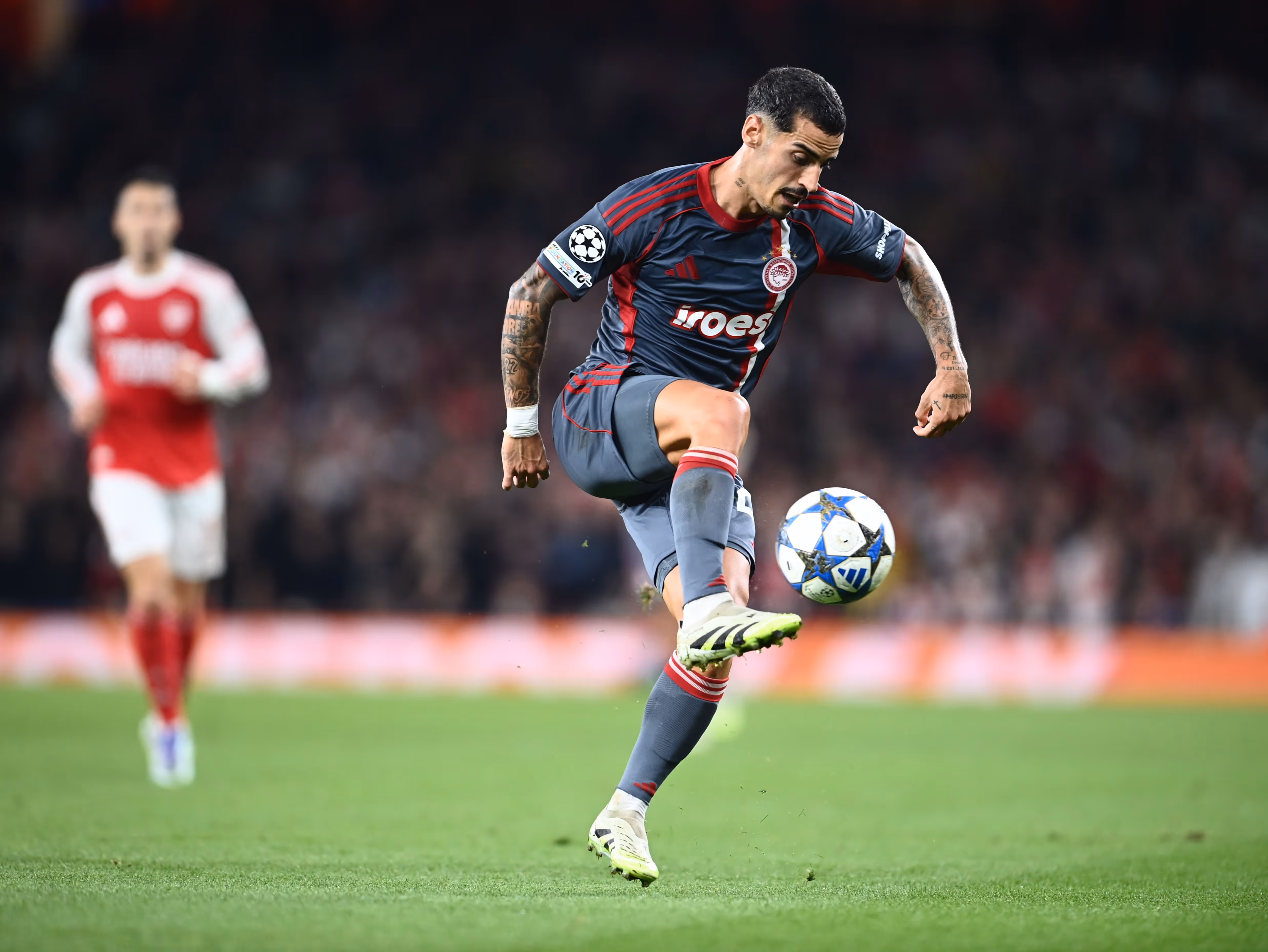 Soccer player in gray and red uniform controlling a ball mid-air during a match with a blurred opponent in red and white in the background.