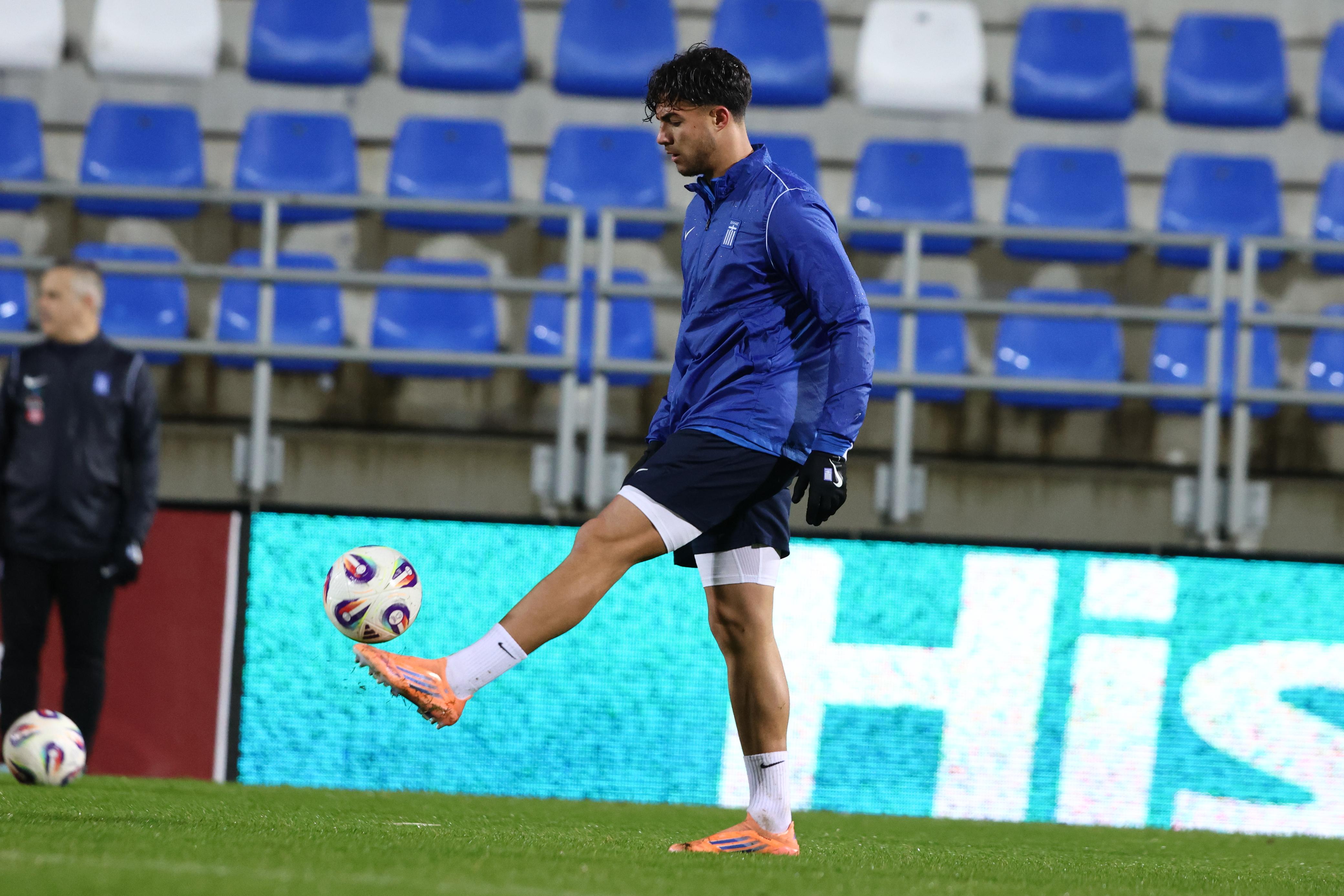 Soccer player in blue jacket and orange cleats juggling a ball on a grass field with empty blue and white stadium seats in the background.