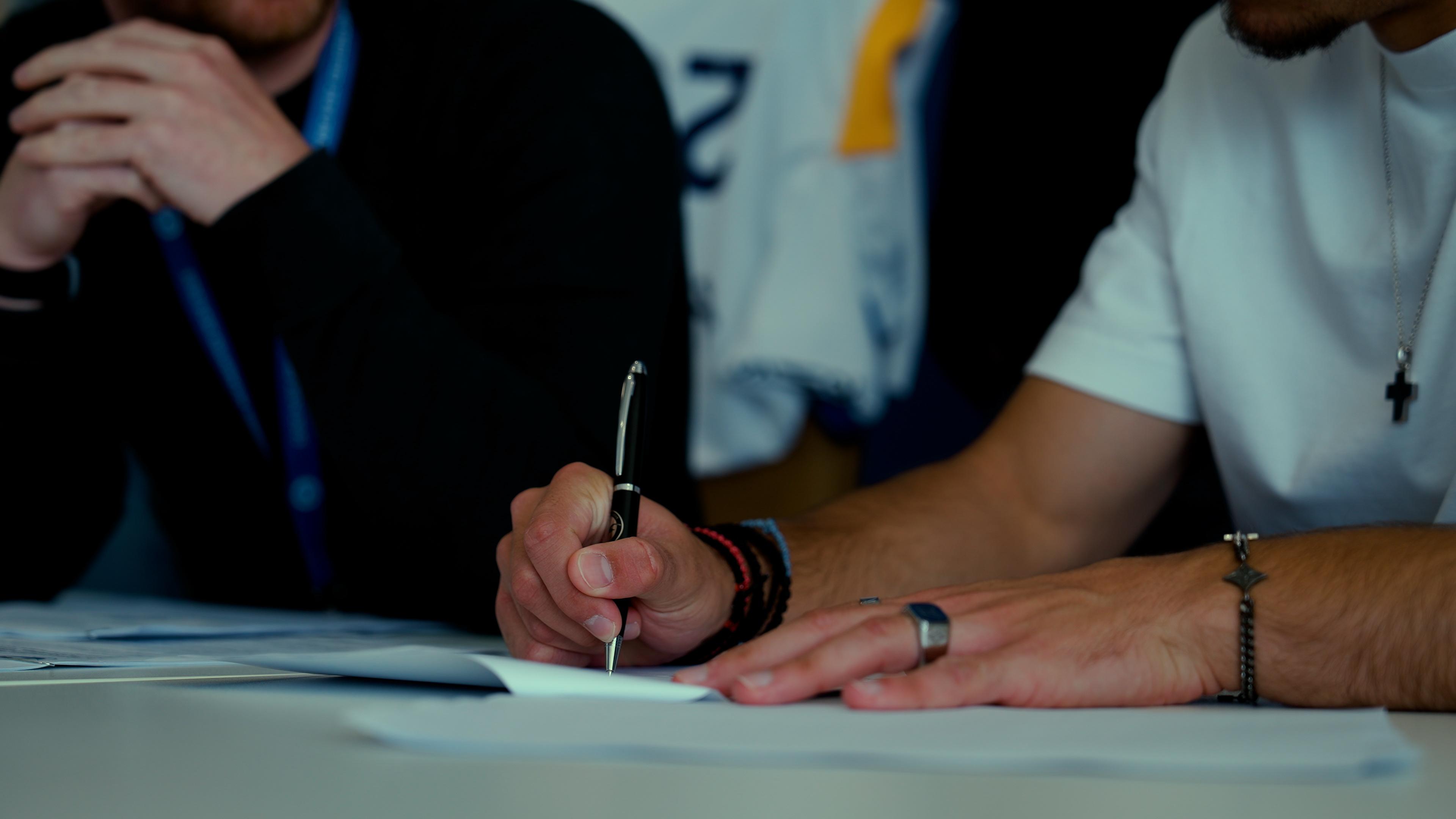 Close-up of two men at a table, one signing a document with a pen while the other watches.