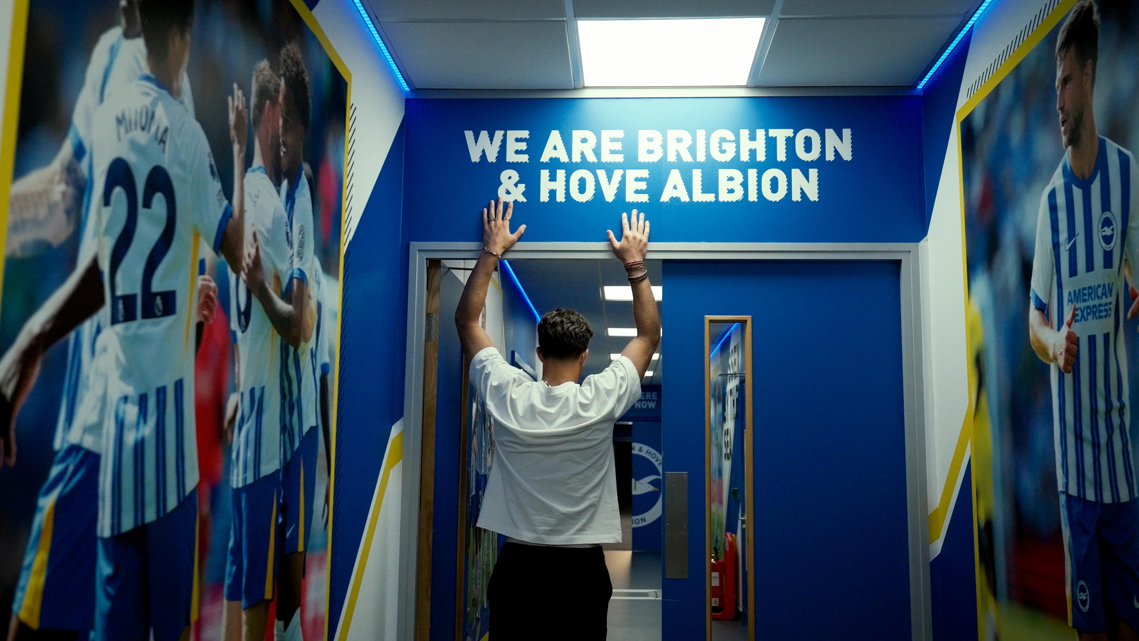 Person facing a blue door with their hands raised under a sign reading 'We Are Brighton & Hove Albion', surrounded by large photos of football players in Brighton & Hove Albion jerseys.