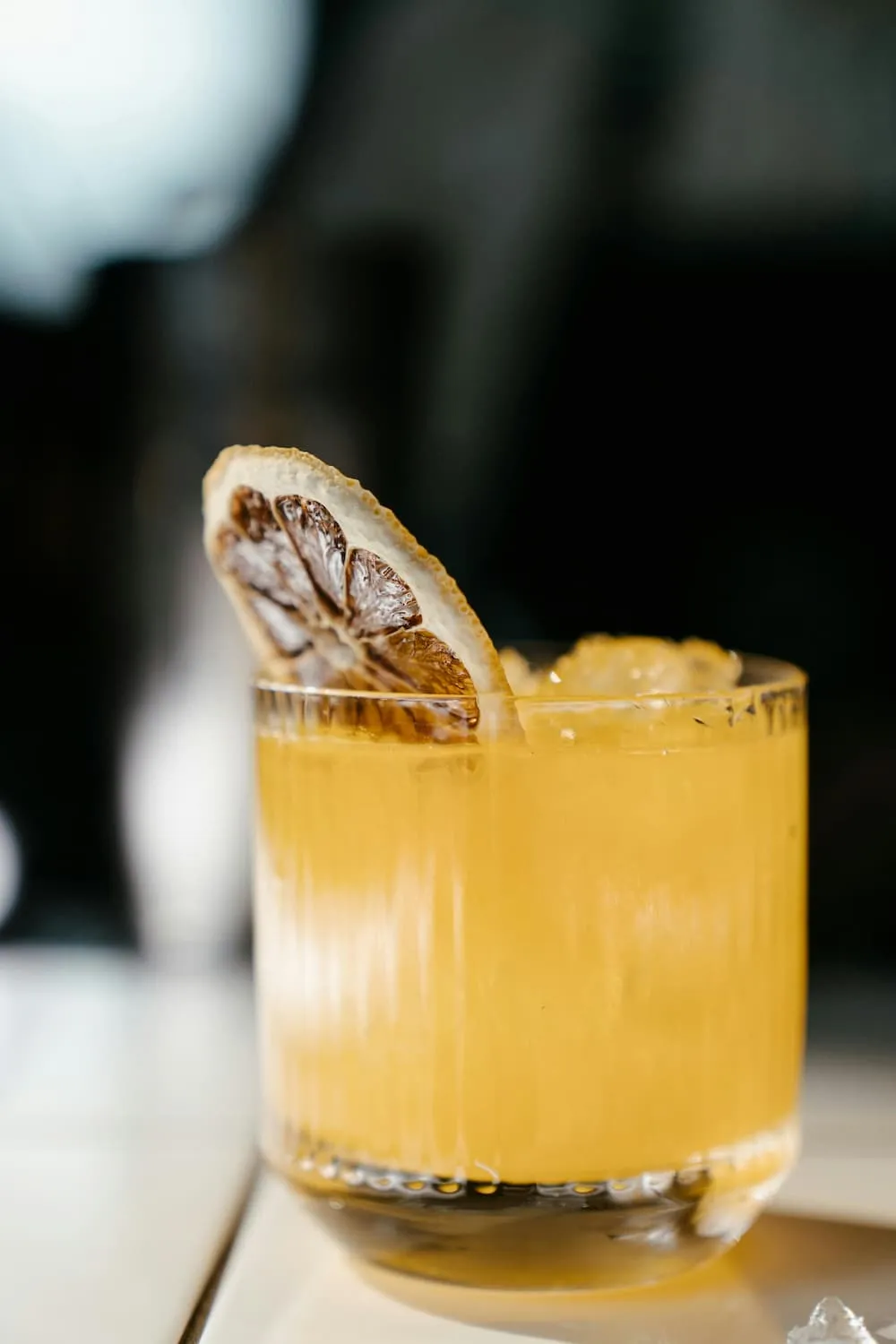 Glass of orange-yellow cocktail with ice cubes and a dried lemon slice garnish on the rim.