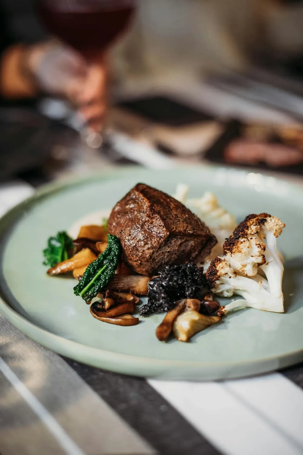 Plate with grilled steak, roasted cauliflower, assorted mushrooms, and leafy greens, with a blurred hand holding a glass of red wine in the background.