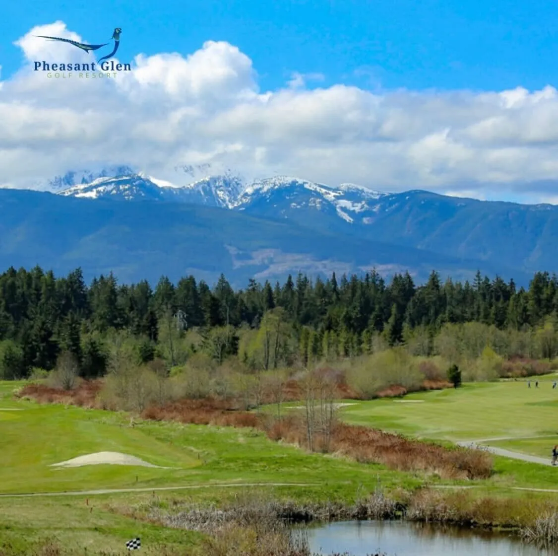 Golf course with green fairways, sand bunker, pond, trees, and snow-capped mountains under a partly cloudy blue sky.