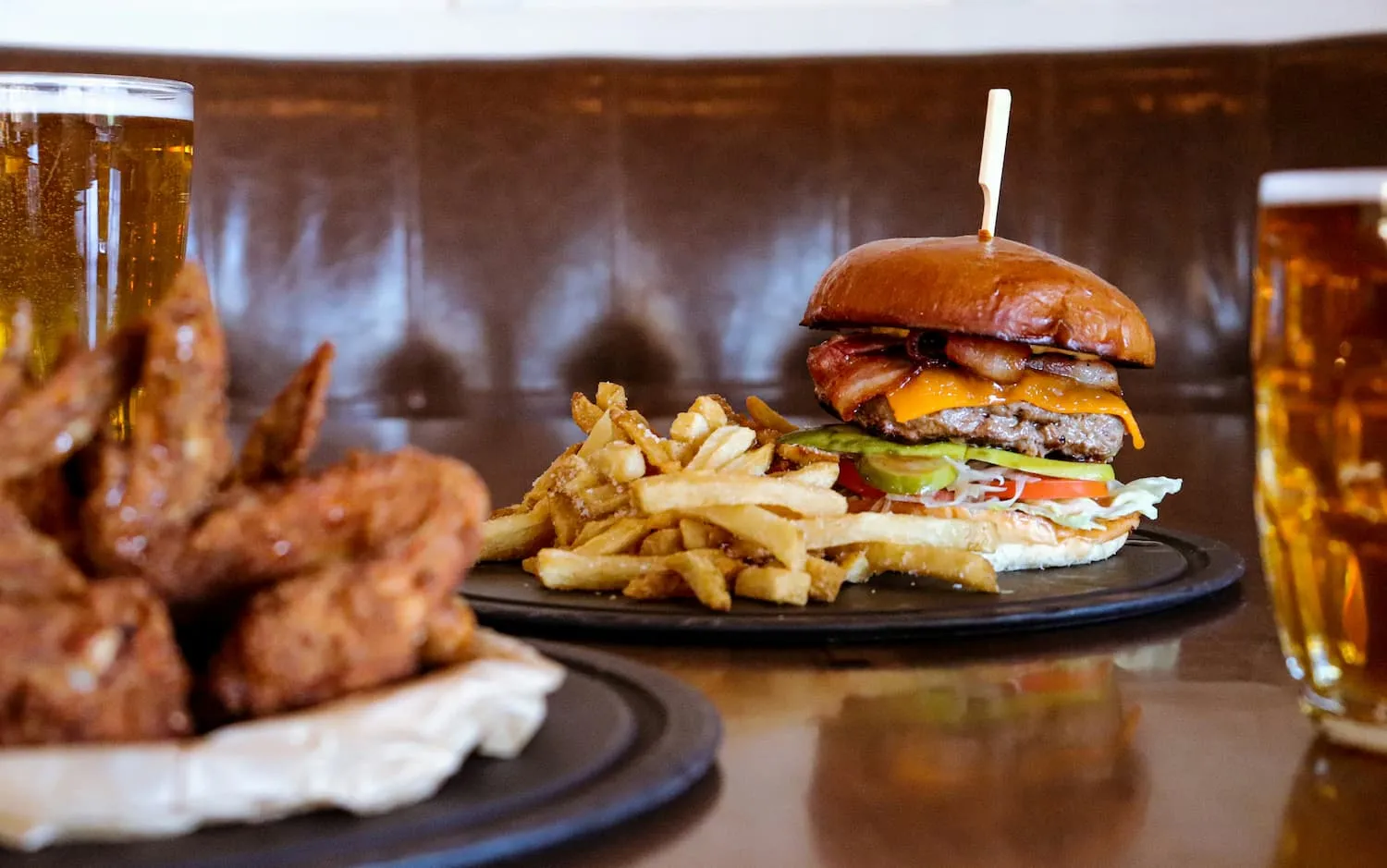 Bacon cheeseburger with lettuce, tomato, pickles, and fries on a black plate, with chicken wings and glasses of beer in the background.