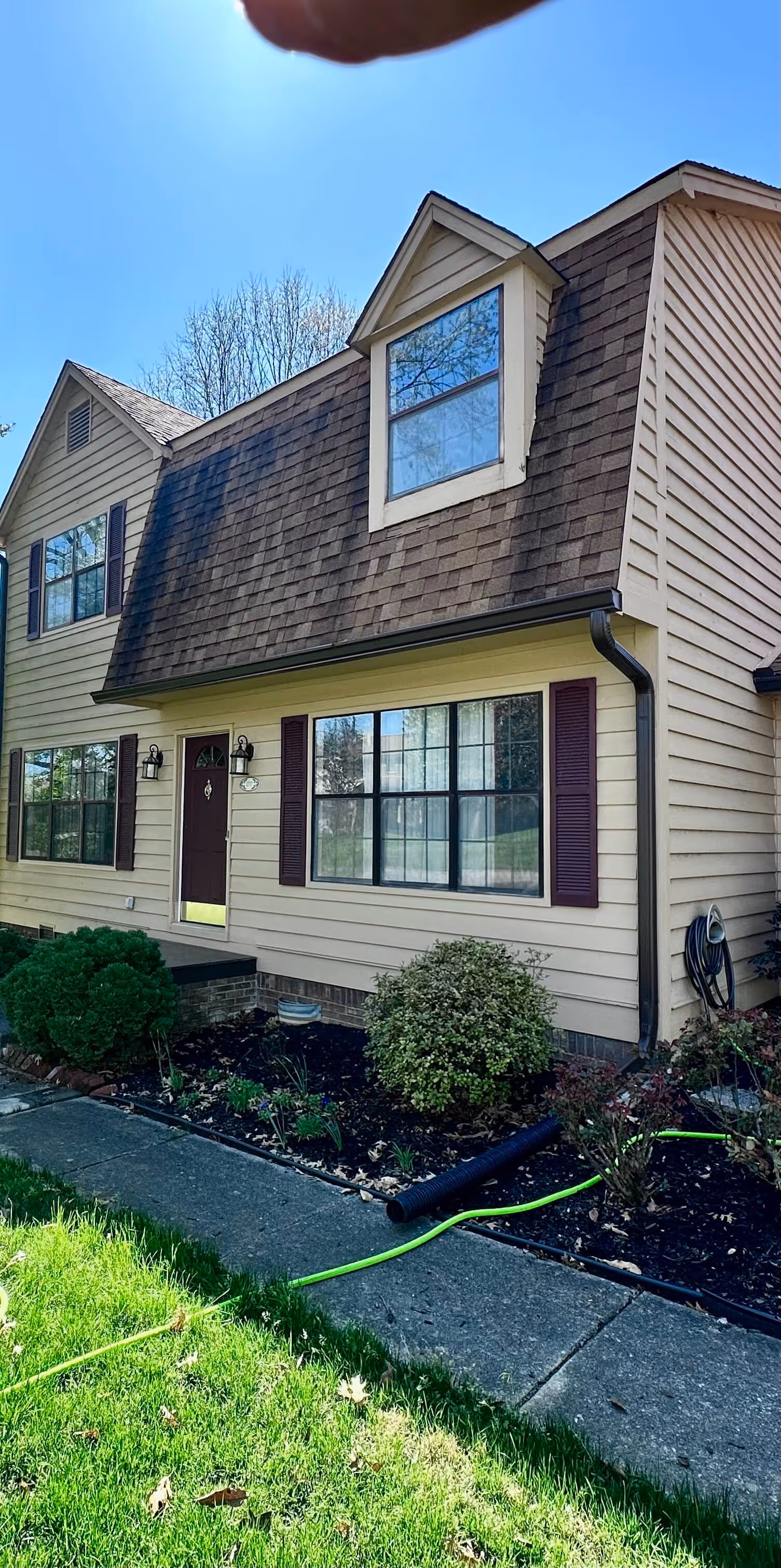 Two-story beige house with brown shingle roof, front door with lights, and garden with bushes 