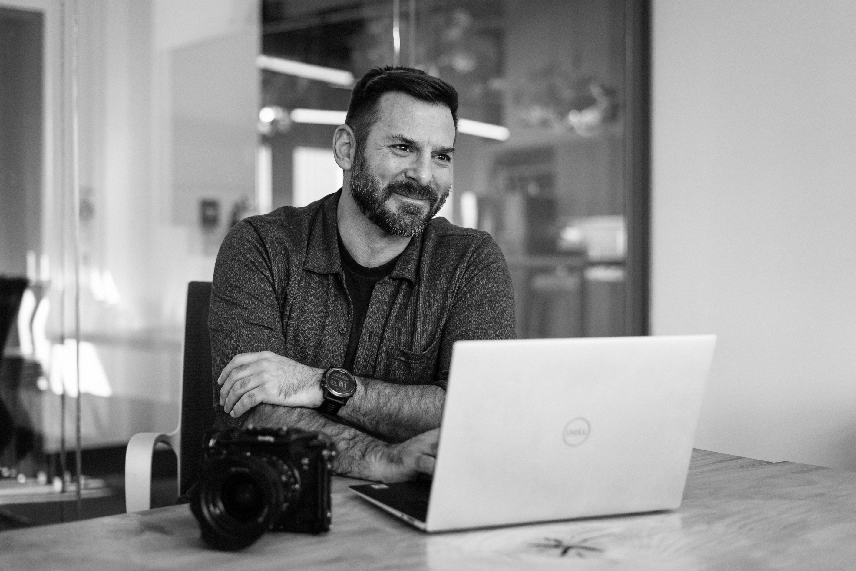 Smiling man with beard sitting at a table with a laptop and camera in a modern office.