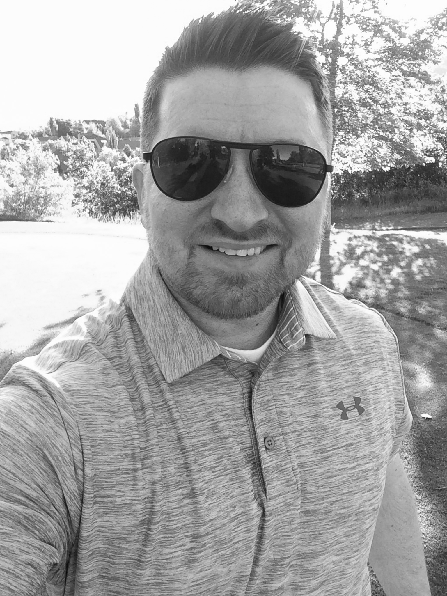 Man wearing sunglasses and a gray Under Armour shirt smiling outdoors on a sunny day.
