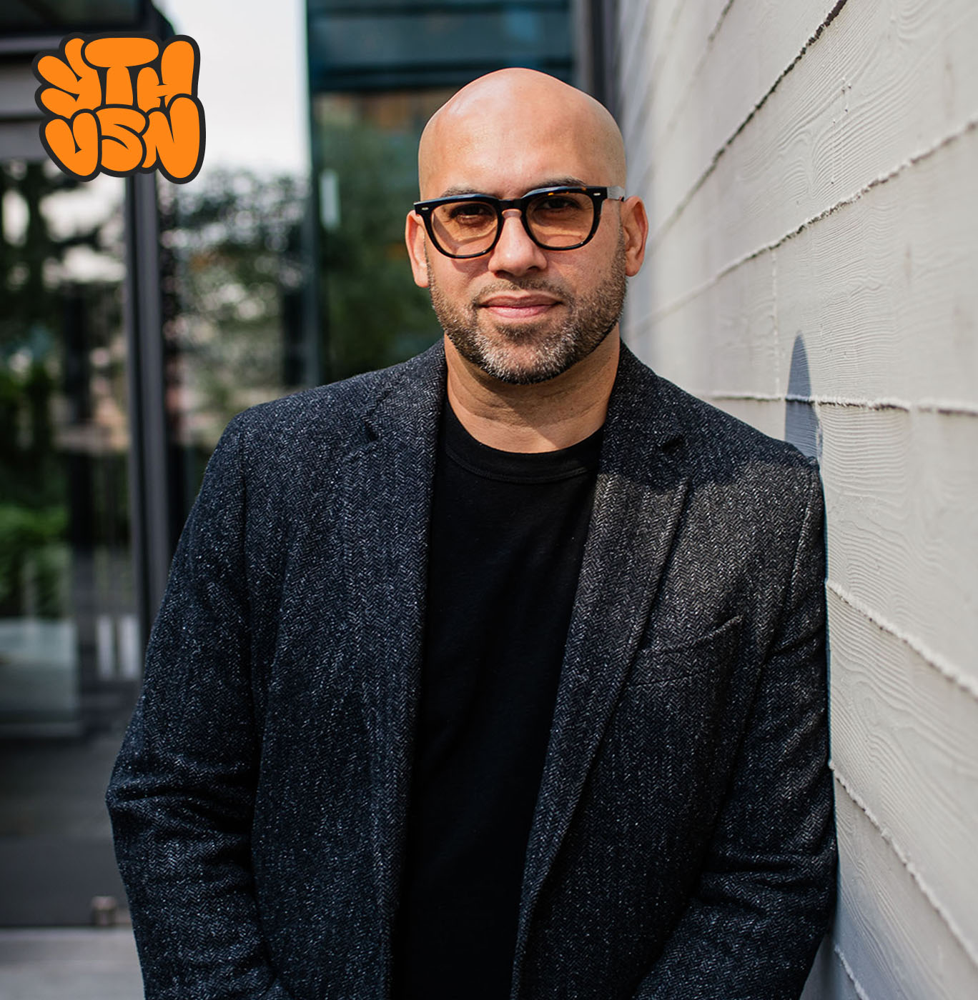 Bald man with glasses and a trimmed beard wearing a black shirt and gray blazer leaning against a white wall outdoors.