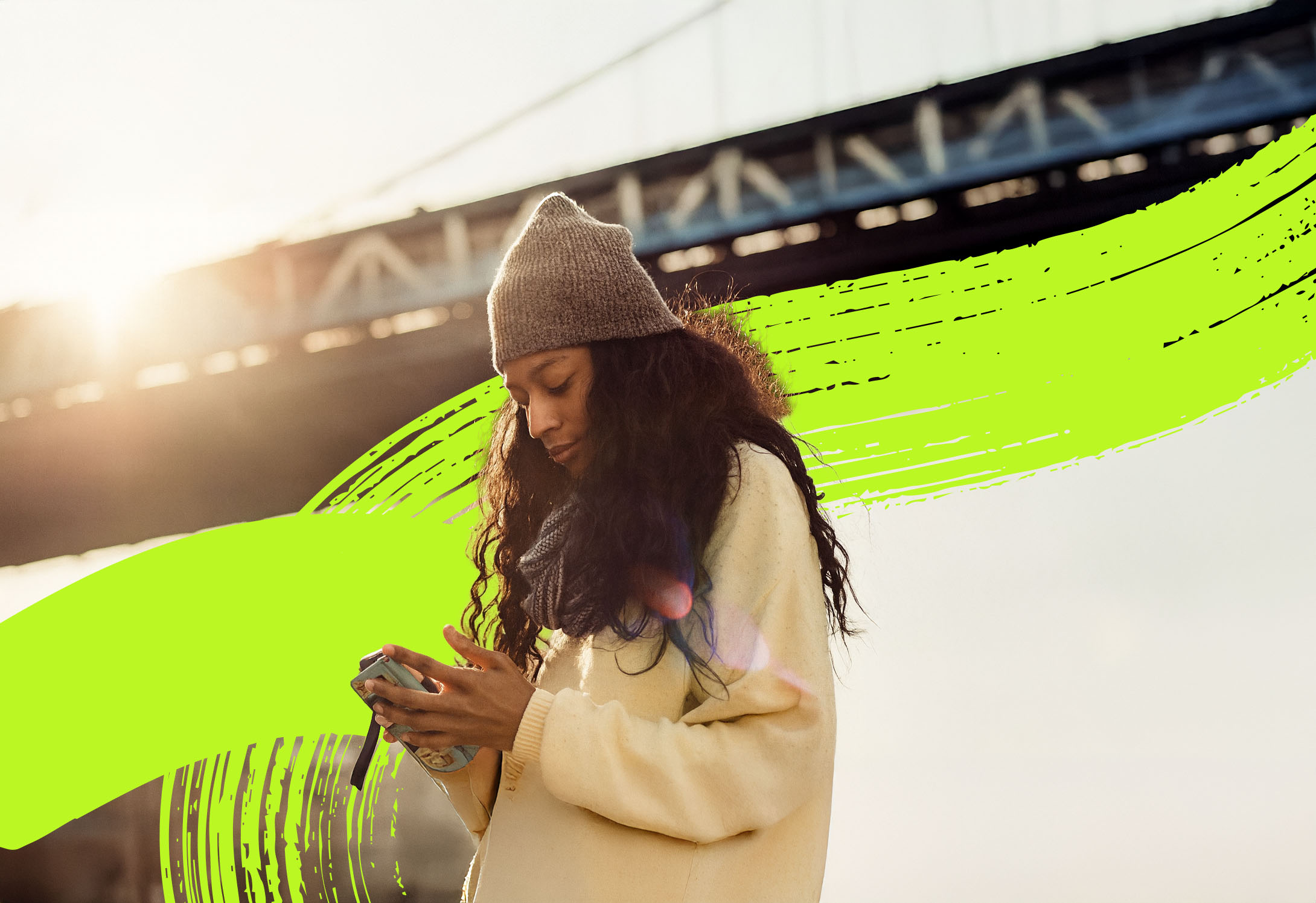 Woman wearing a gray knit hat and cream coat, focused on her phone with a bridge and sunlight in the background.