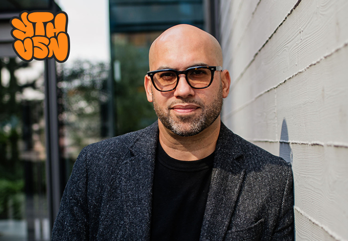 Bald man with glasses and trimmed beard wearing a dark blazer and black shirt standing against a textured white wall outdoors.