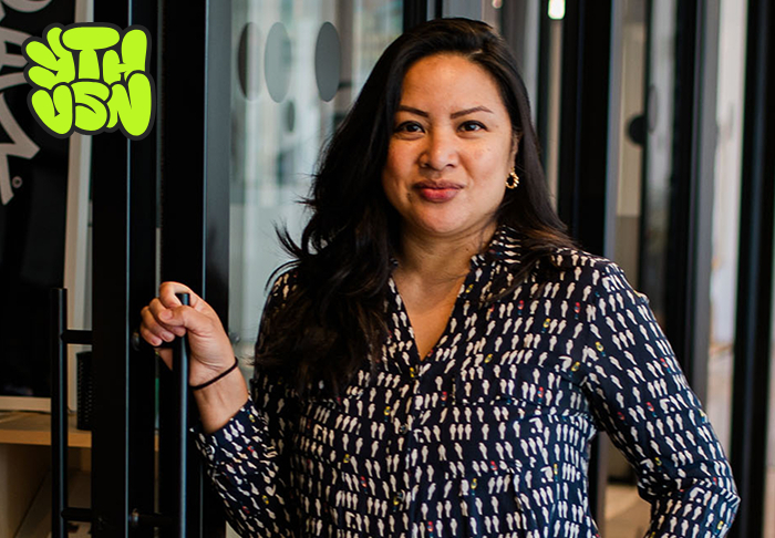 Smiling woman with long dark hair wearing a patterned shirt standing indoors near glass doors.