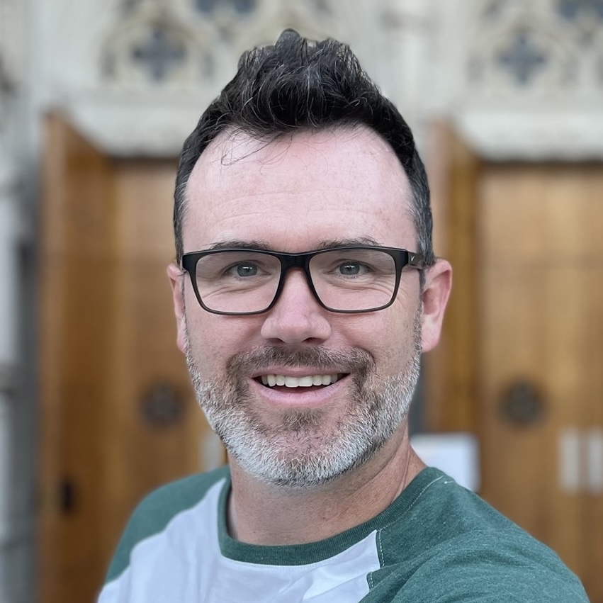 Smiling man with short dark hair, a salt-and-pepper beard, and black-framed glasses wearing a green and white shirt.