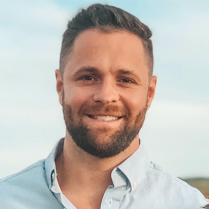Smiling man with short dark hair and beard wearing a light blue button-up shirt against a blue sky background.