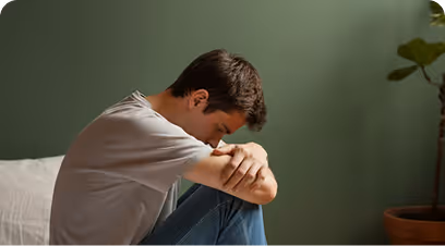 Joven sentado con los brazos apoyados en las rodillas y la cabeza inclinada hacia abajo en una habitación con pared verde.