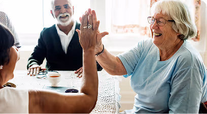 Dos personas mayores sonrientes dándose un choque de manos en una mesa mientras un hombre observa y sonríe.
