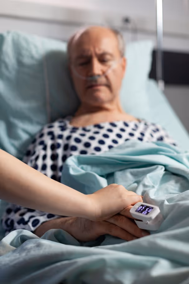 Paciente mayor en cama de hospital con oxígeno nasal mientras alguien sostiene su mano con un oxímetro de pulso.