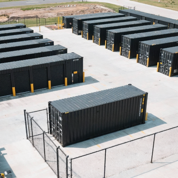 Aerial view of multiple black shipping containers arranged in rows on a concrete lot enclosed by a chain-link fence.