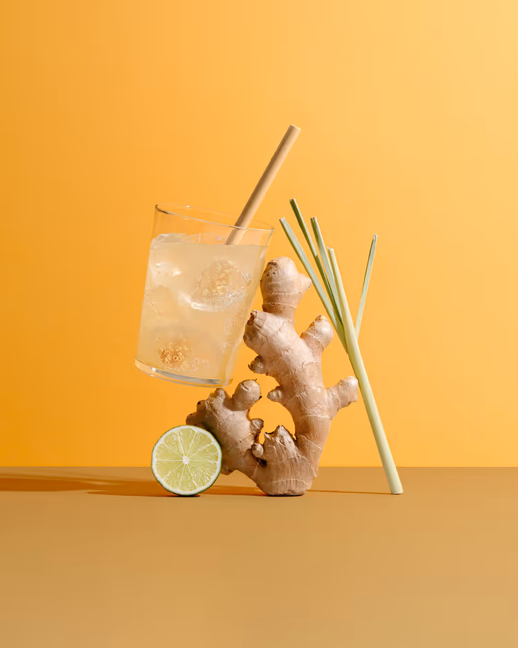 Glass of iced lemon ginger drink with a bamboo straw, fresh ginger root, lemongrass stalks, and a lime half on a brown surface against an orange background.