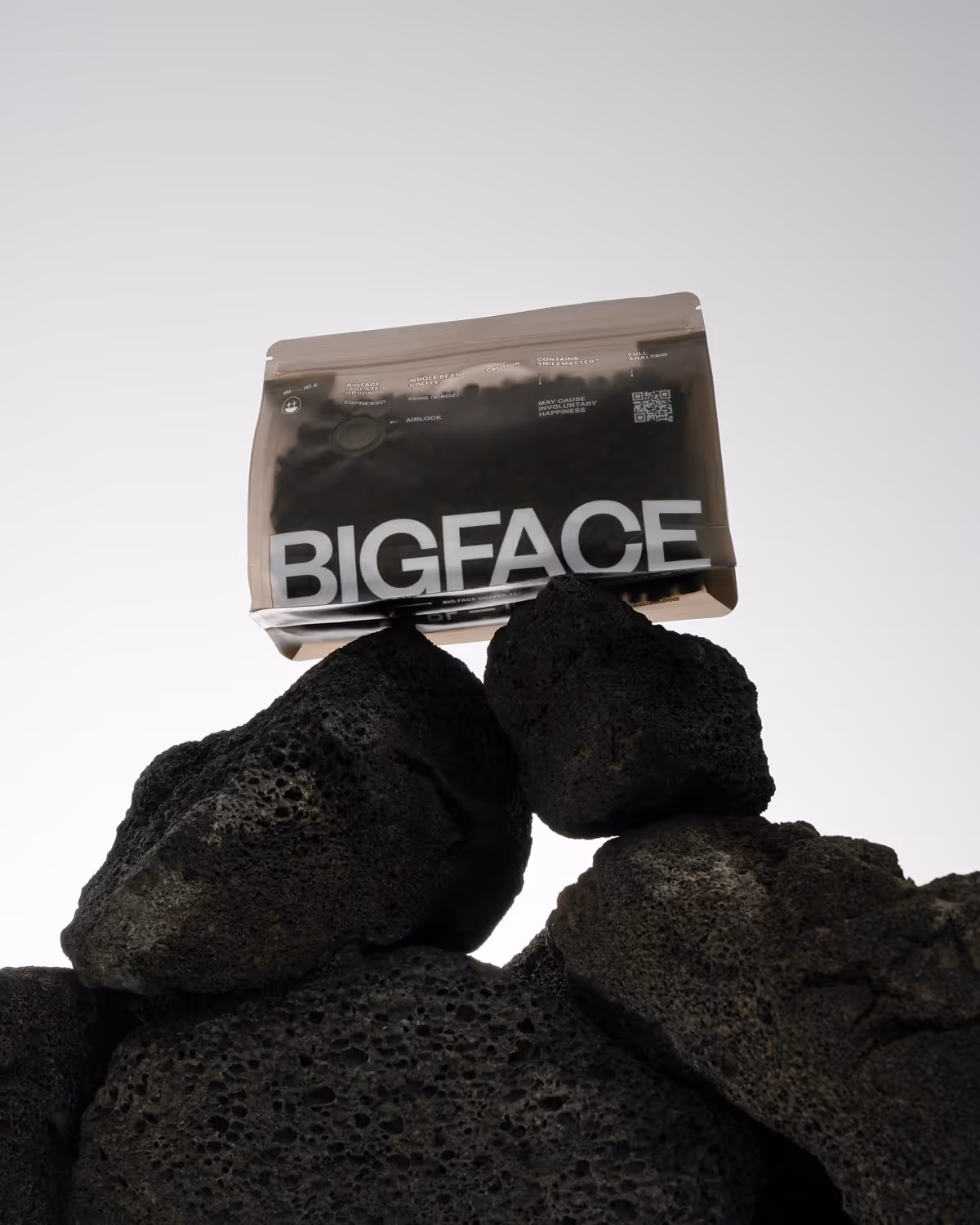 Bag of BIGFACE whole bean coffee resting on stacked black volcanic rocks against a clear sky.