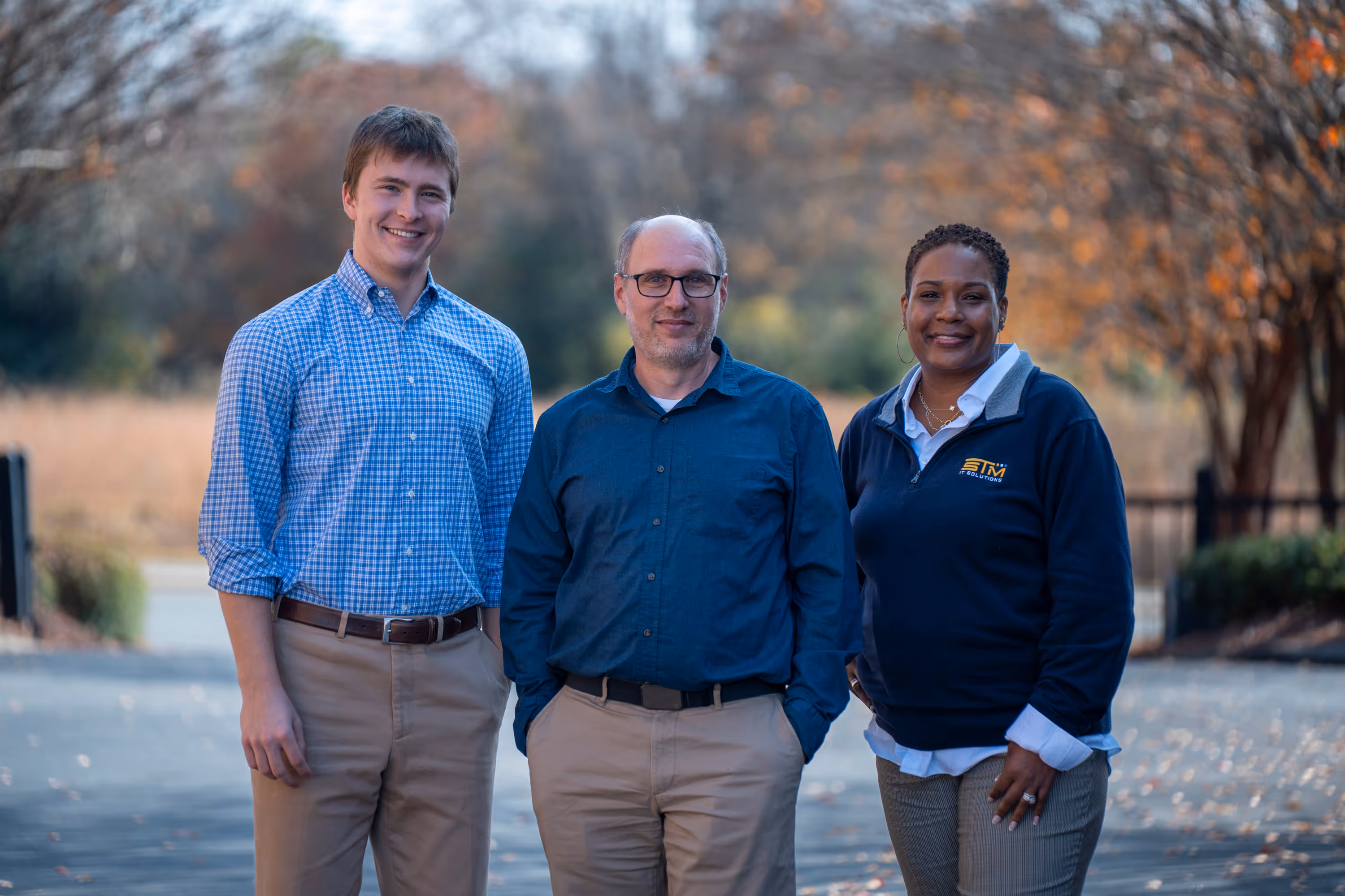 Three adults standing outdoors on a paved path with autumn trees in the background, smiling at the camera.