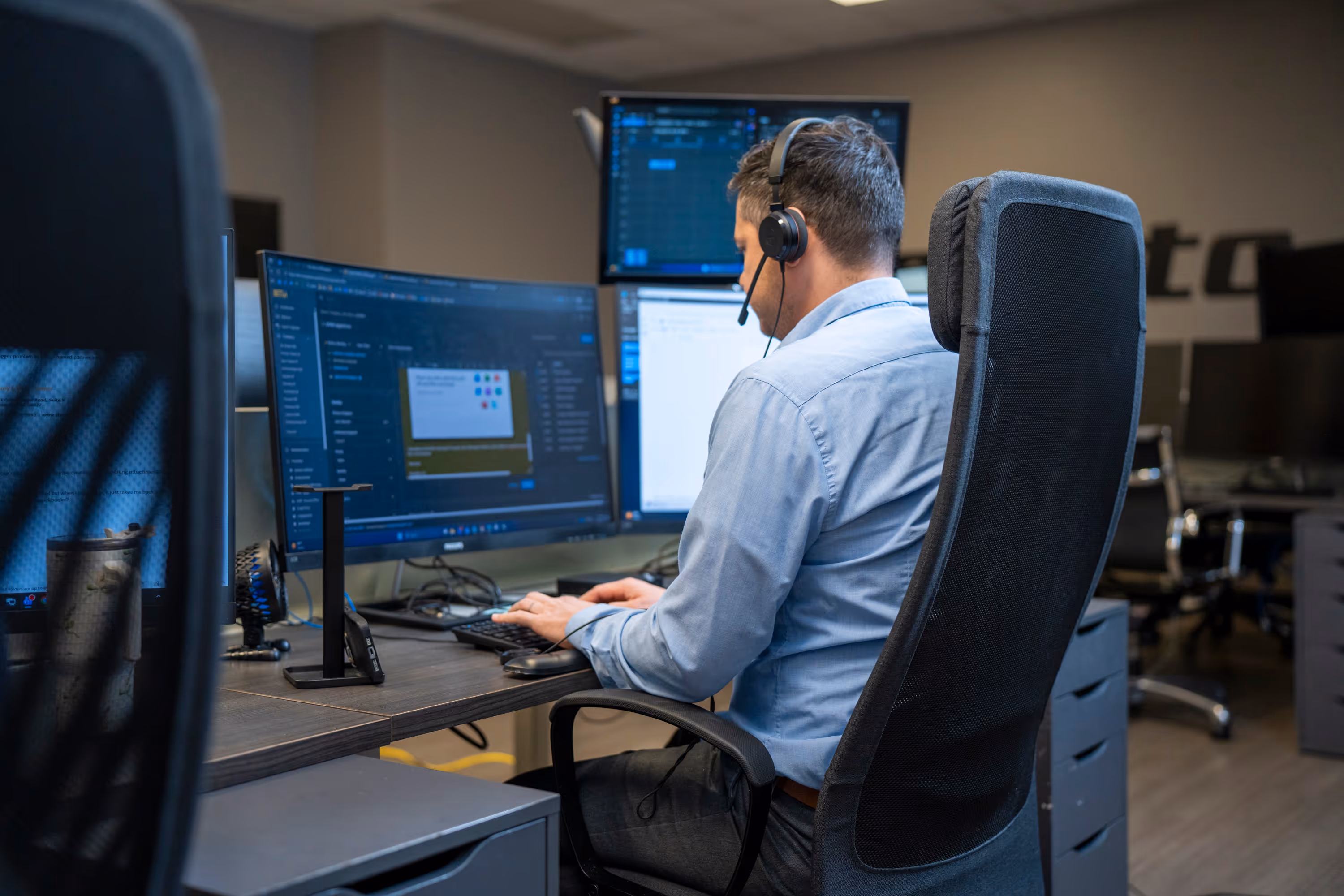 Man wearing a headset working on multiple computer monitors in an office.