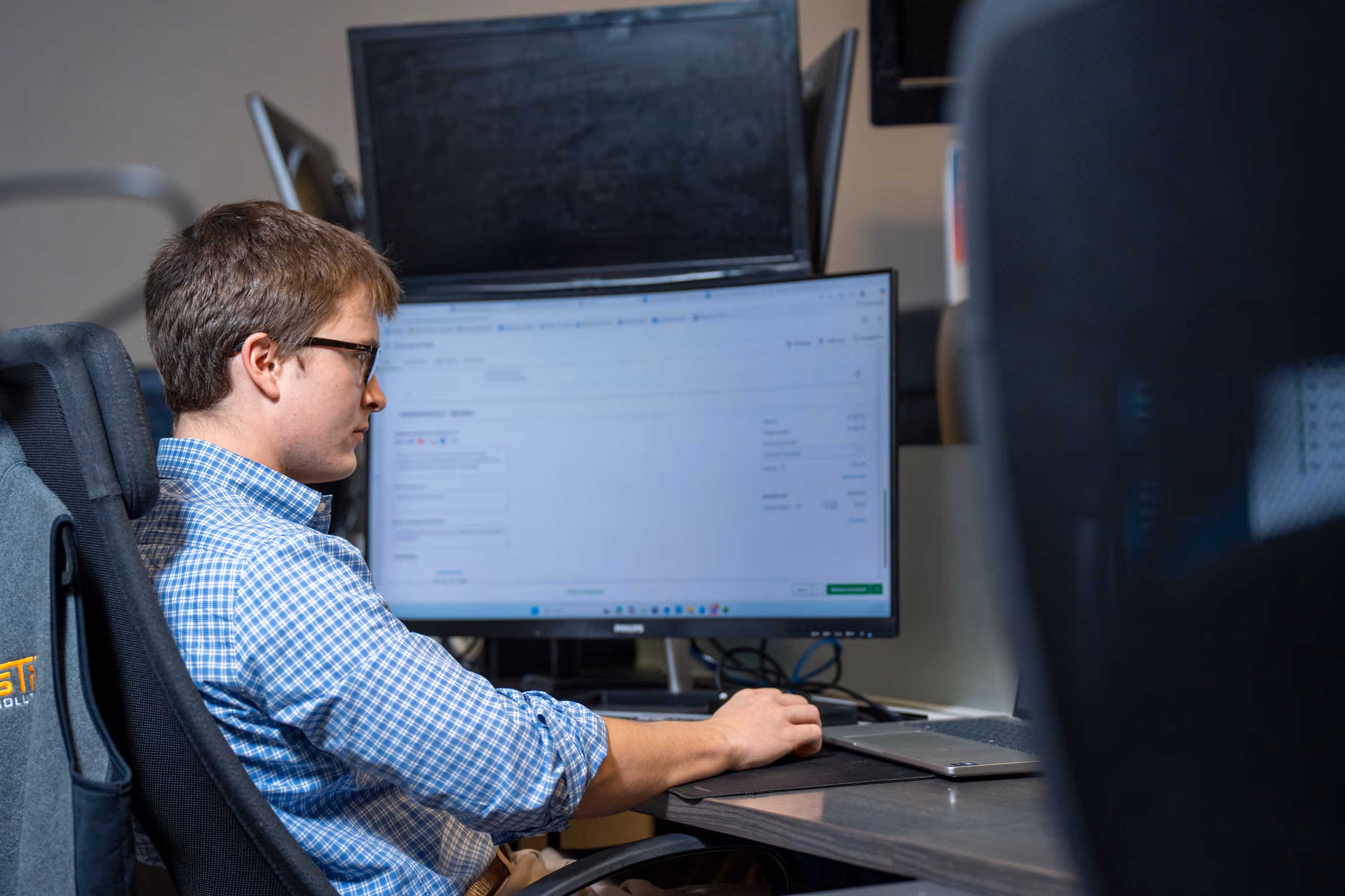 Young man in glasses and a blue checkered shirt working on a laptop at a desk with a large monitor displaying a webpage.