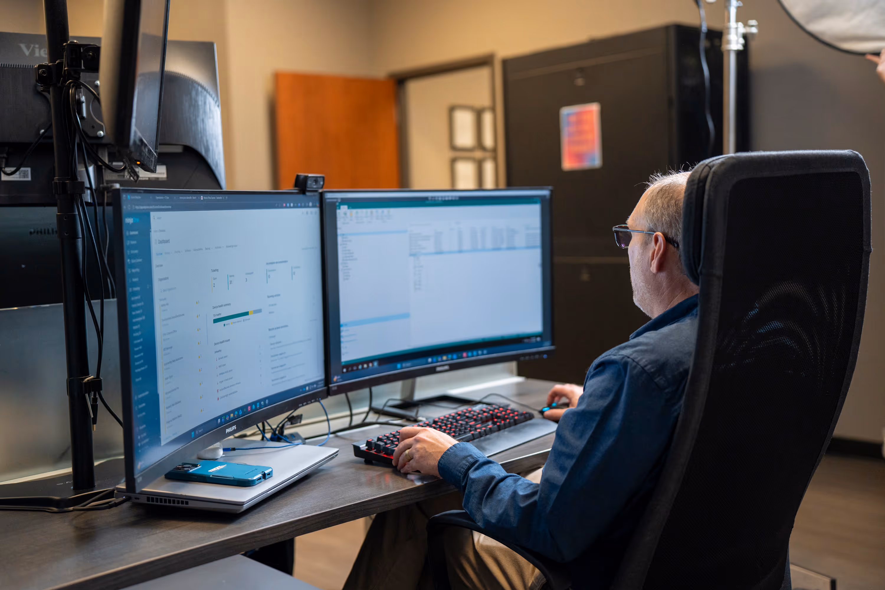 Man in glasses working at a desk with two large computer monitors displaying dashboards and data.