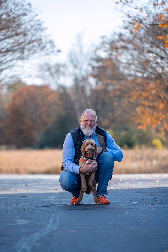 Smiling man with a white beard squatting on a road while holding a small brown dog wearing an orange harness, with autumn trees in the background.