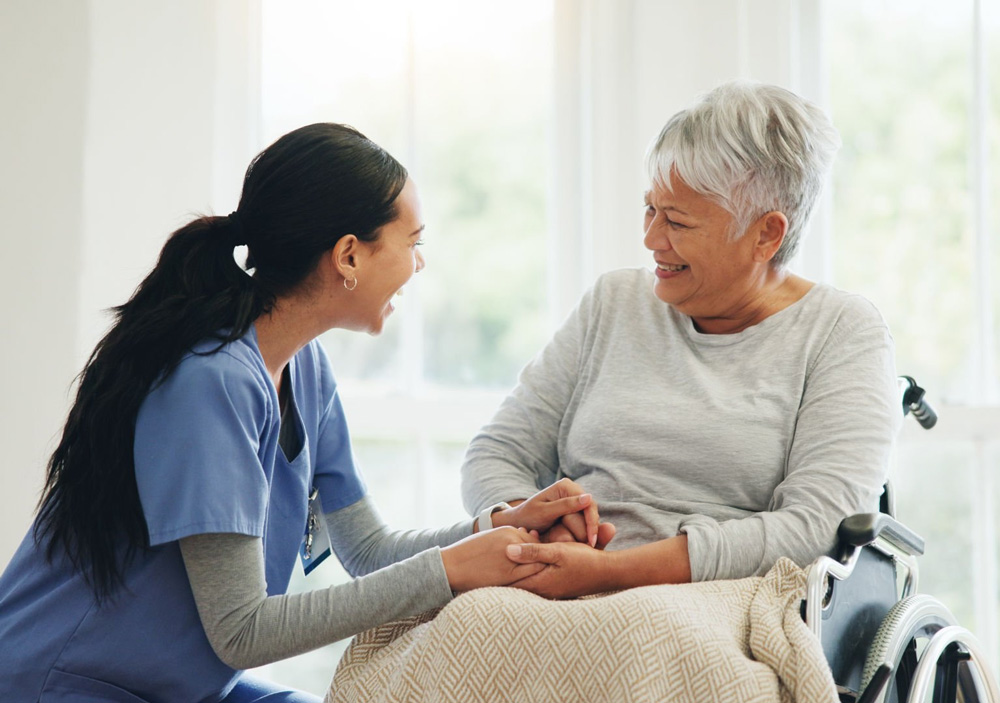 Caregiver in blue scrubs holding hands and smiling with elderly woman in a wheelchair covered with a blanket.