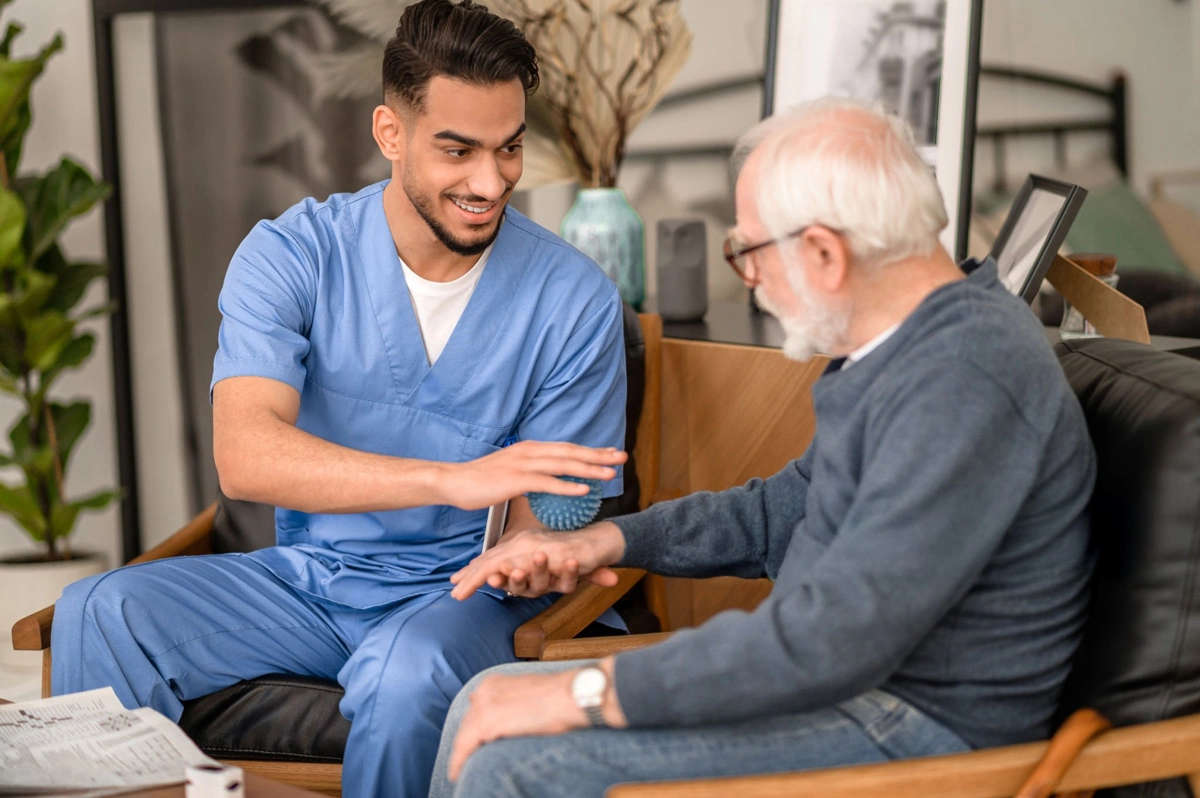 Young male therapist in blue scrubs using a spiky massage ball on an elderly man's wrist during a therapy session.