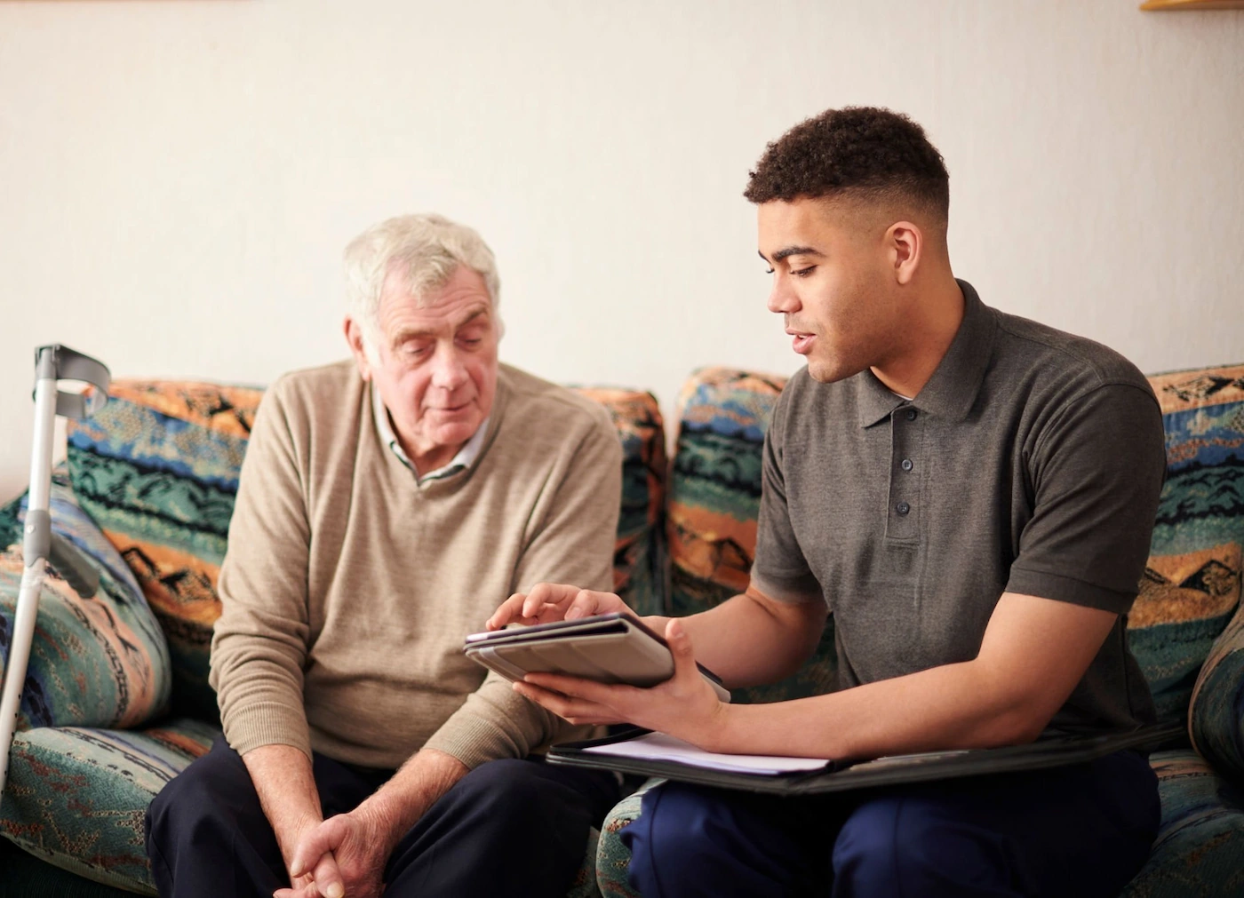 Young man showing a tablet to an elderly man sitting together on a patterned couch.