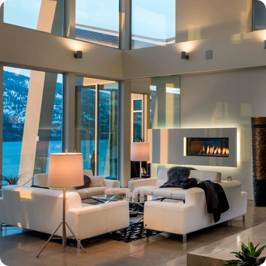 Modern living room with white sofas, a glass coffee table, floor lamps, a wall-mounted fireplace, and large windows showing a lake and mountains.