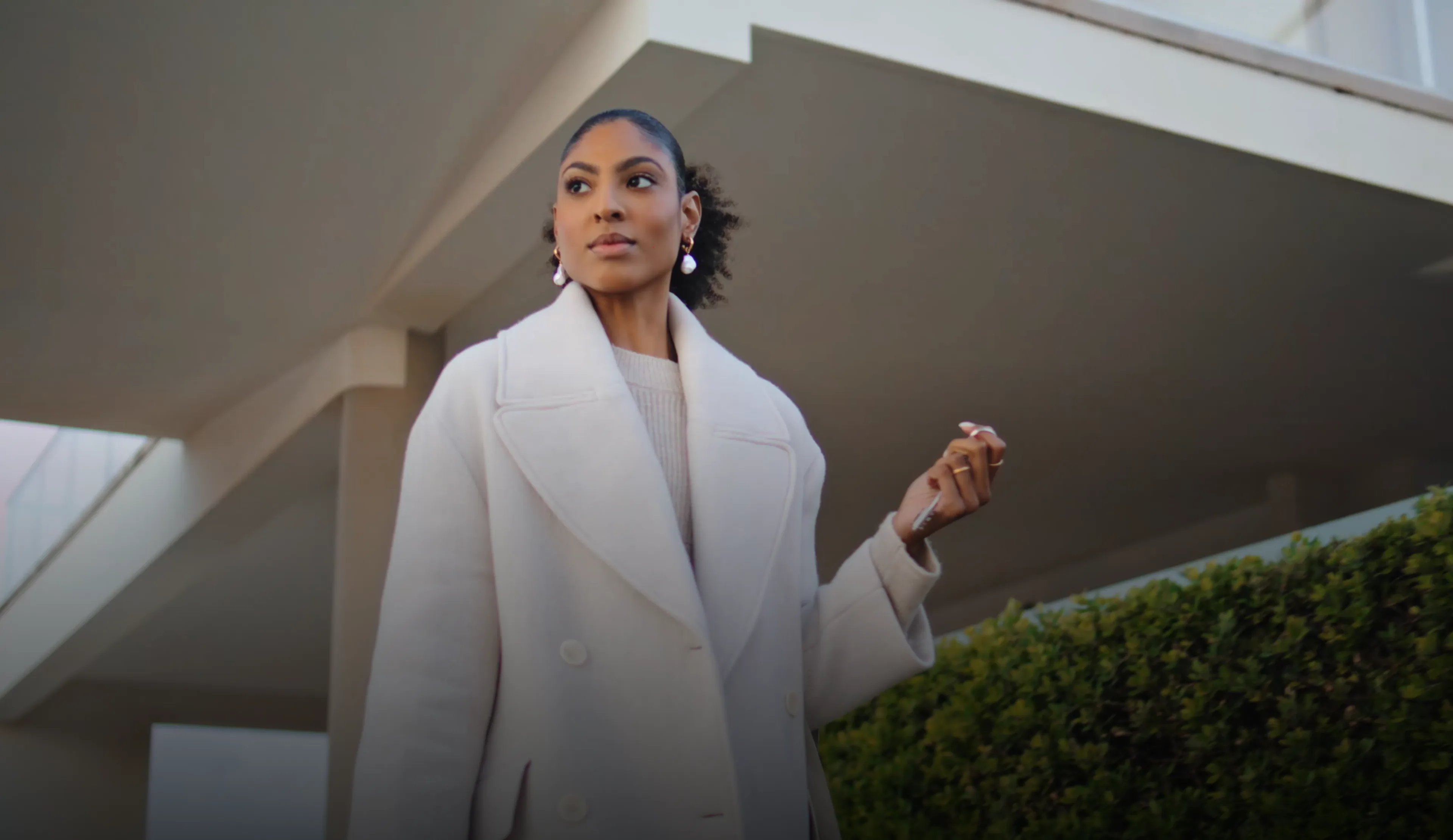 Woman with pulled-back hair wearing a white coat, standing outdoors near modern building architecture and greenery.