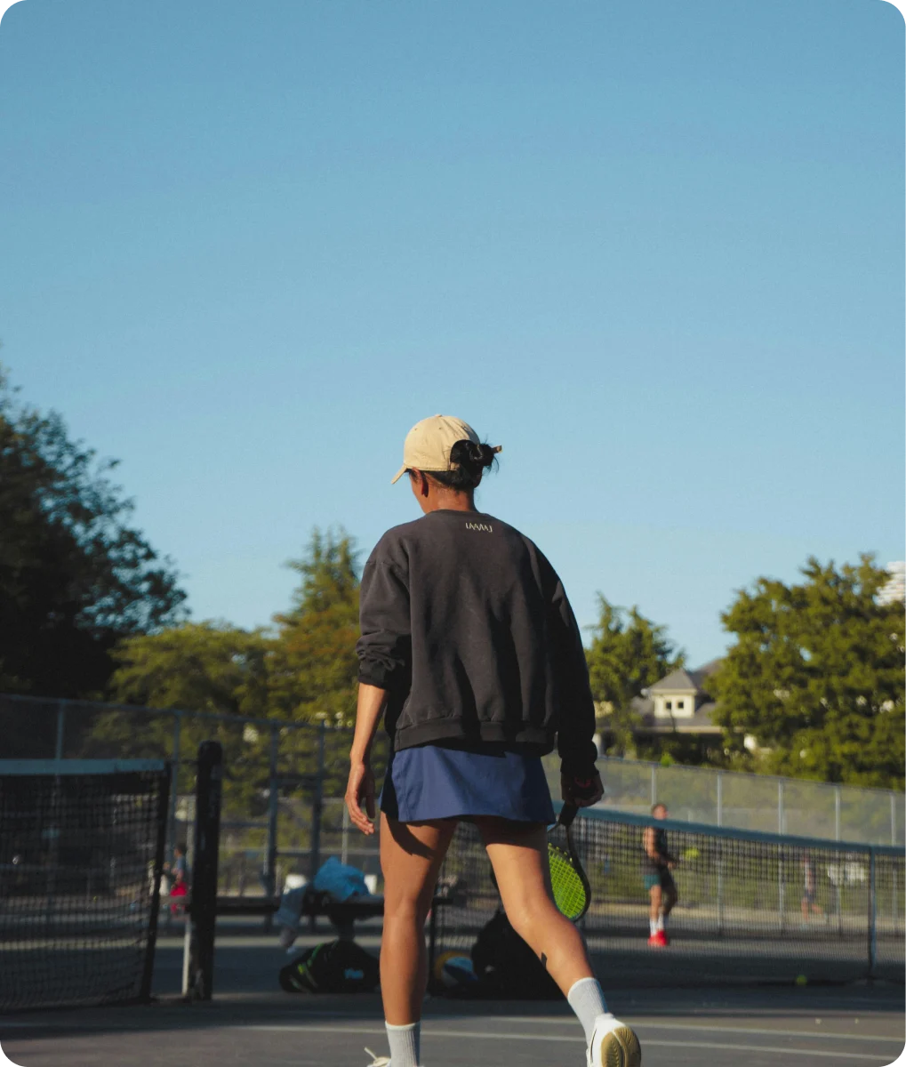 Person wearing a beige cap, black sweatshirt, and blue shorts walking on an outdoor tennis court holding a tennis racquet.