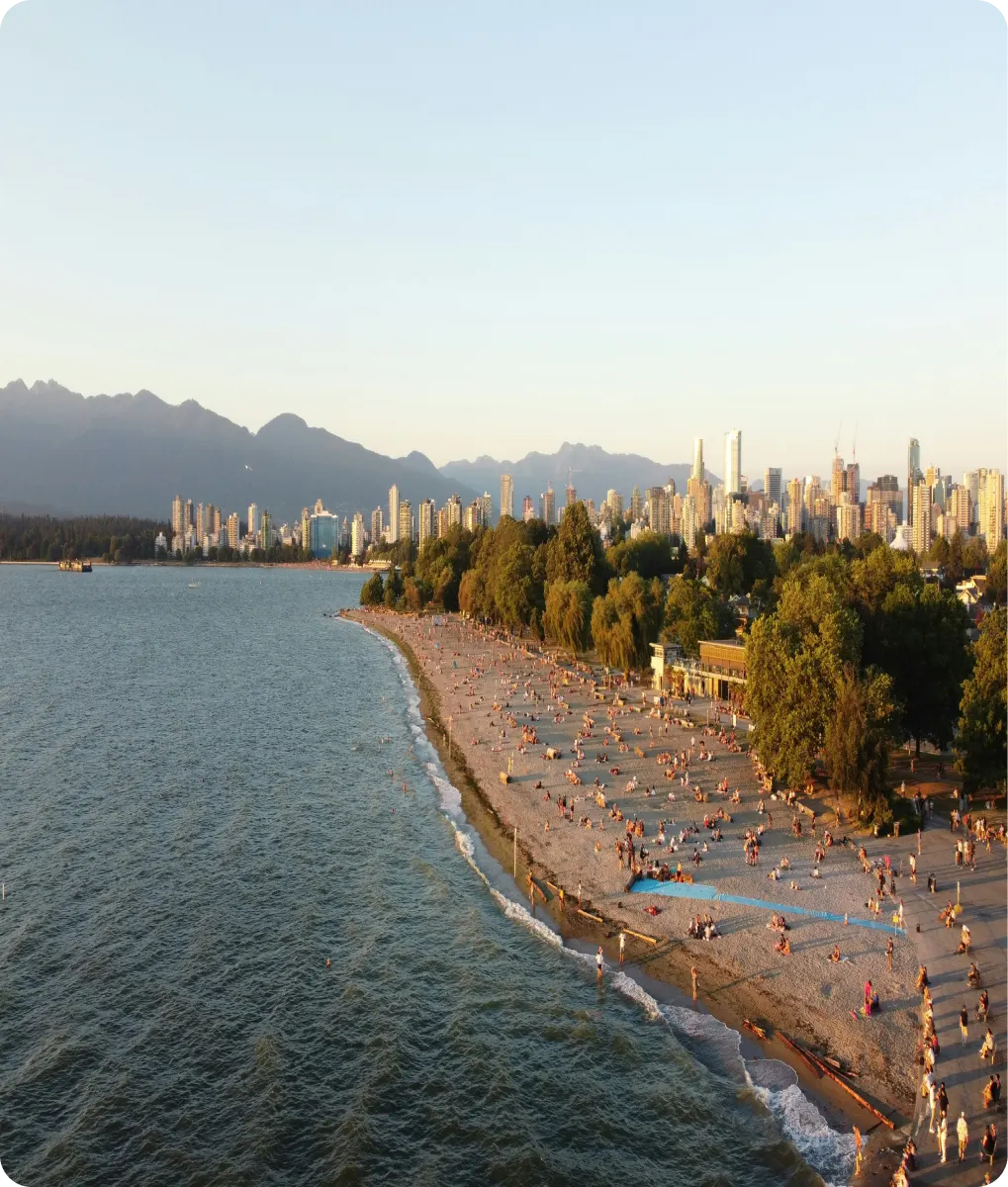 A crowded sandy beach with people enjoying the sun, bordered by trees and a city skyline with mountains in the background.