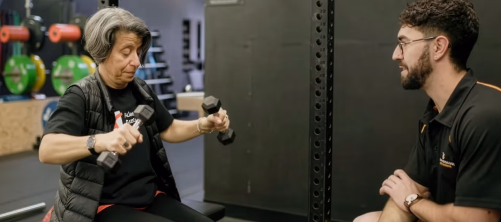Older woman lifting dumbbells while a exercise physiologists in sportswear observes and coaches her in a gym setting.