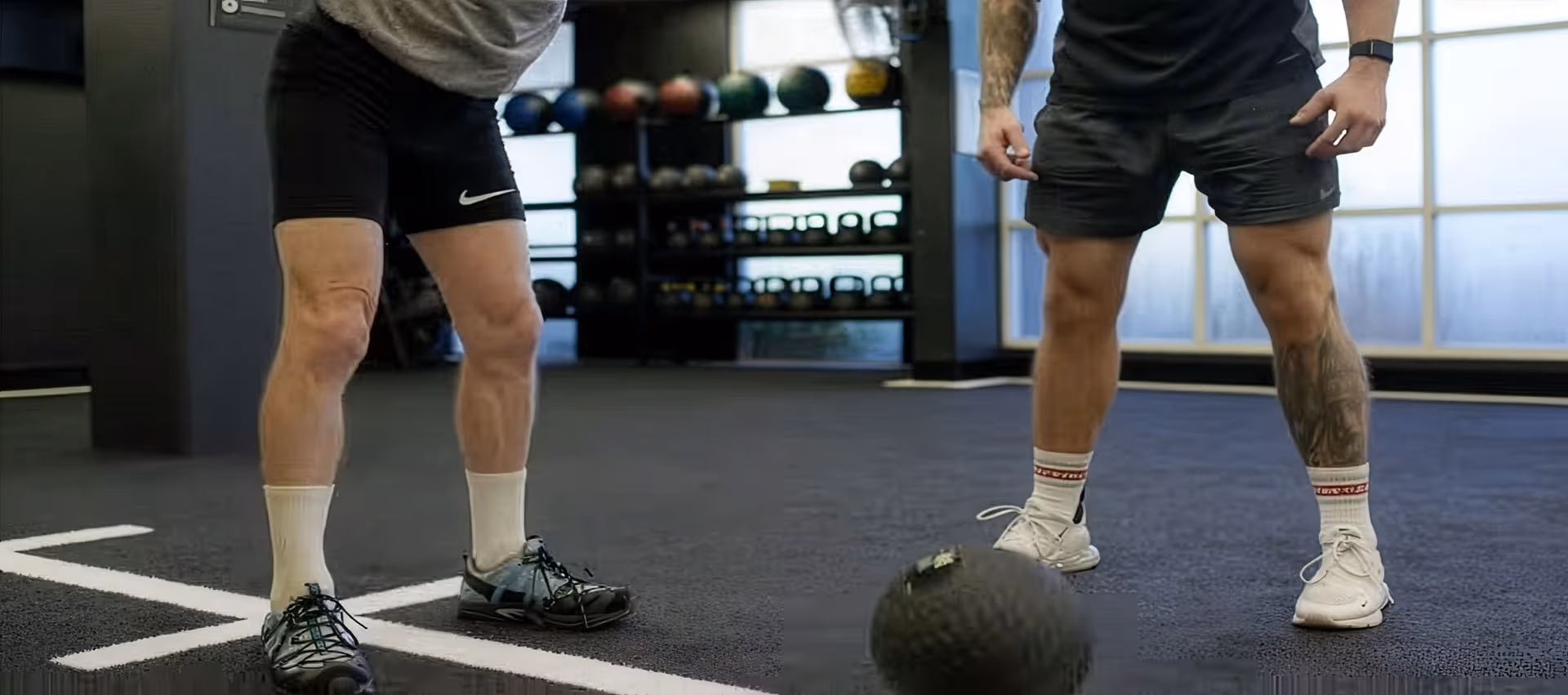 Two men in shorts and athletic shoes standing on a gym floor facing a medicine ball.