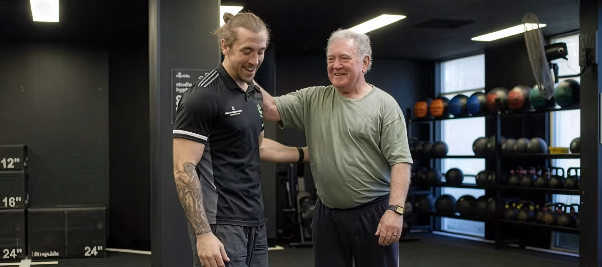 Older man smiling with his hand on the shoulder of a male exercise physiologist inside a gym.