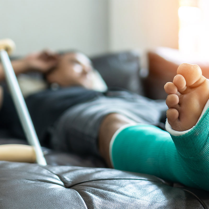 Person lying on a couch with a green leg cast and a crutch beside them.