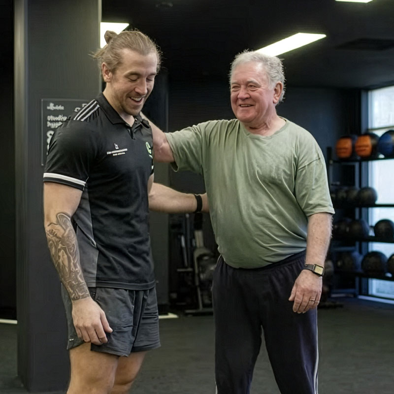 Younger male exercise physiologist  with tattoos smiling and interacting with an older man in workout clothes inside a gym.