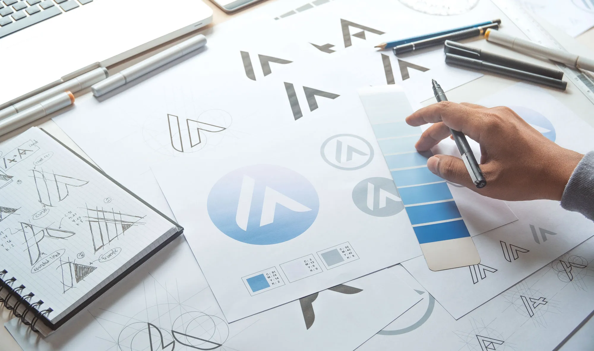 Hand holding a pen over blue color swatches beside sheets with logo designs and sketches on a desk.
