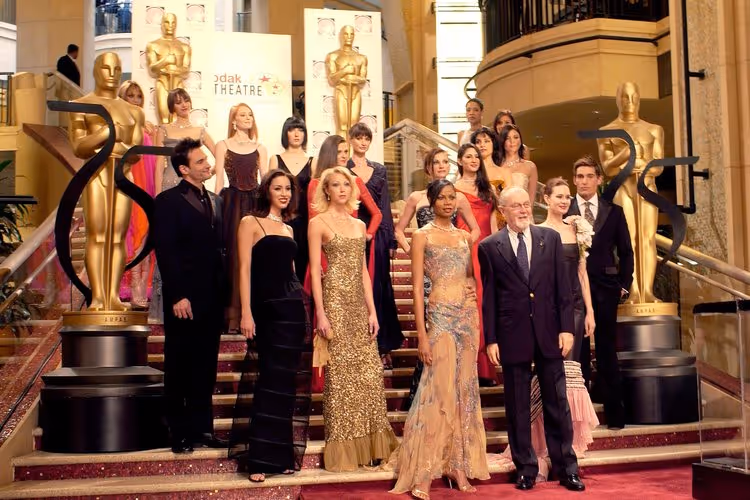 Group of people dressed in formal evening wear posing on grand staircase at Kodak Theatre with large Oscar statues in the background.