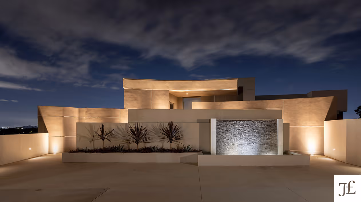 Modern beige concrete house exterior lit at night with a clean driveway and small plants in front.