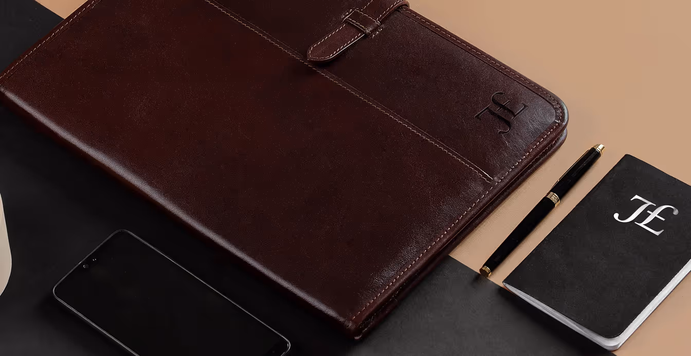 Dark brown leather folio with embossed initials 'JL', next to a black pen and notebook with matching 'JL' initials, and a smartphone on a two-tone black and beige surface.