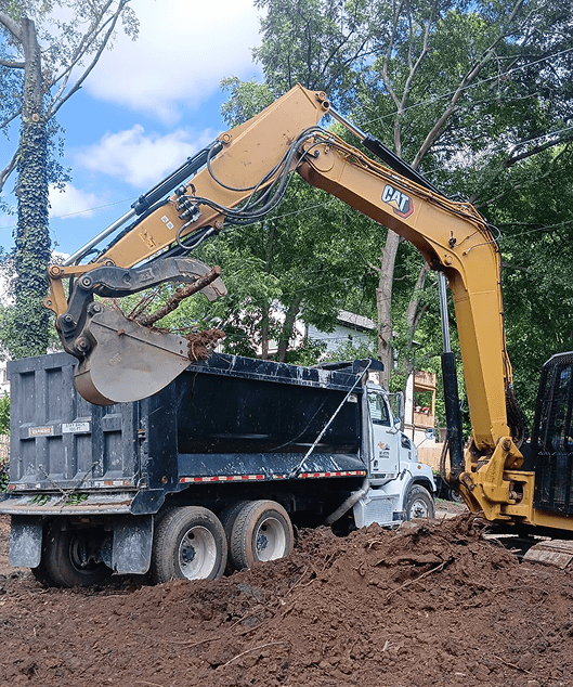 Yellow CAT excavator loading dirt into a black dump truck at a construction site with trees in the background.
