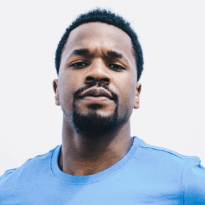 Portrait of a serious young man with short curly hair and beard wearing a blue shirt against a white background.