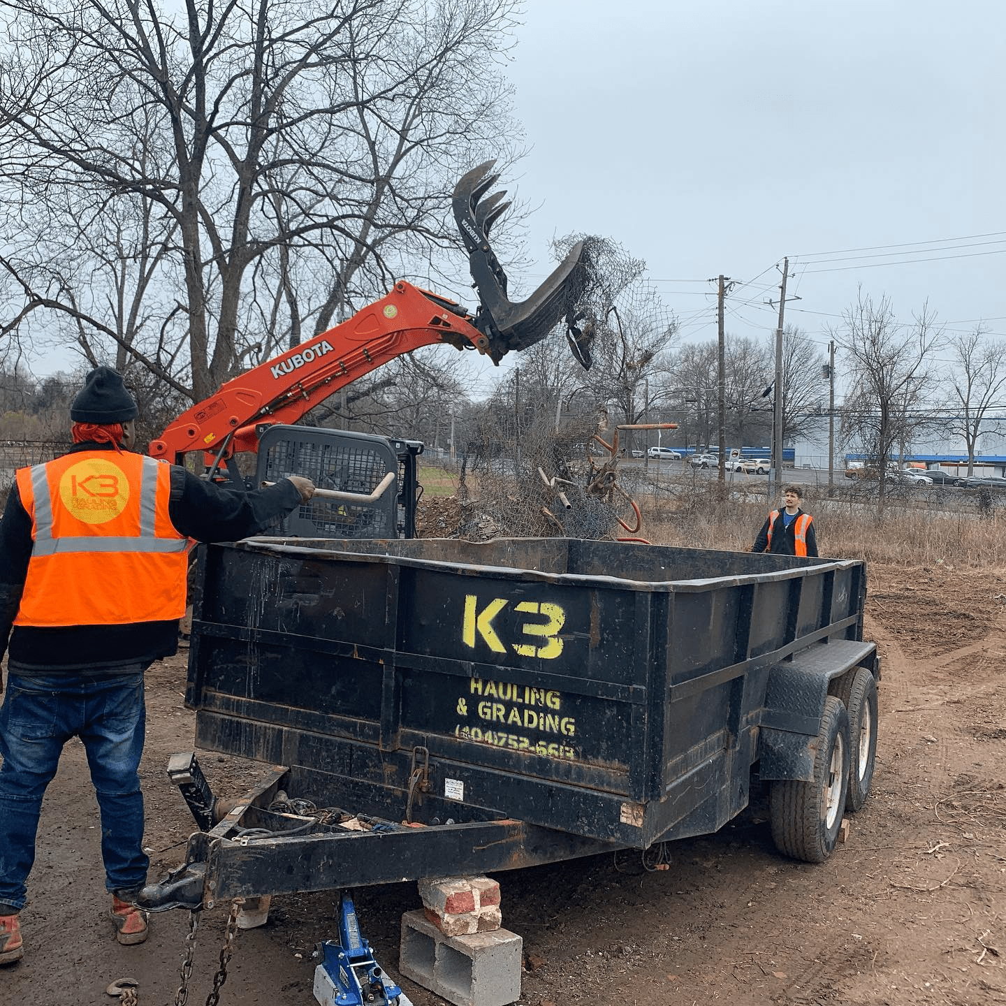 A worker in an orange vest operates a Kubota excavator to load debris into a black K3 Hauling & Grading trailer on a dirt construction site.