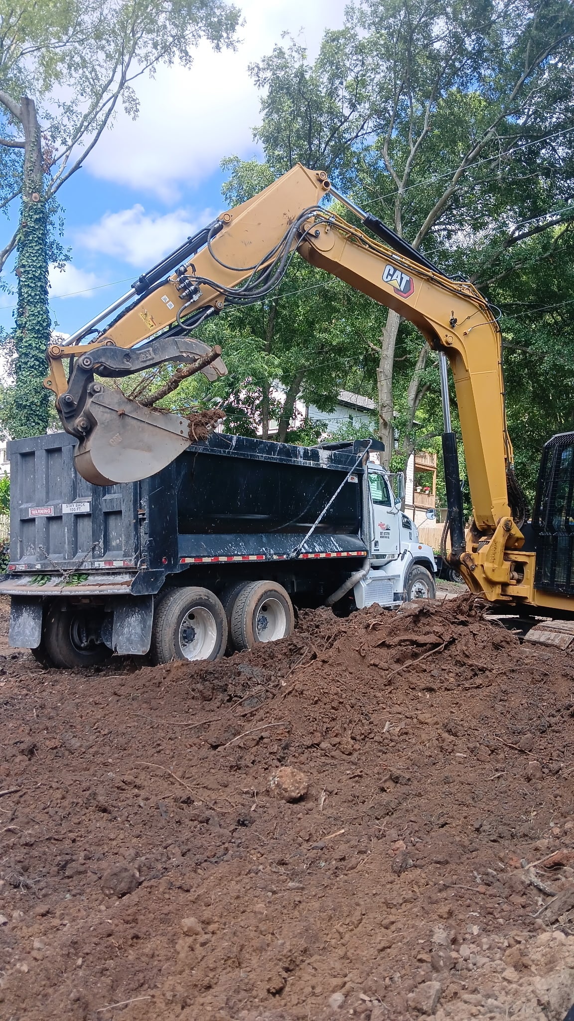 Yellow CAT excavator loading dirt and tree roots into the back of a black dump truck at a construction site surrounded by trees.