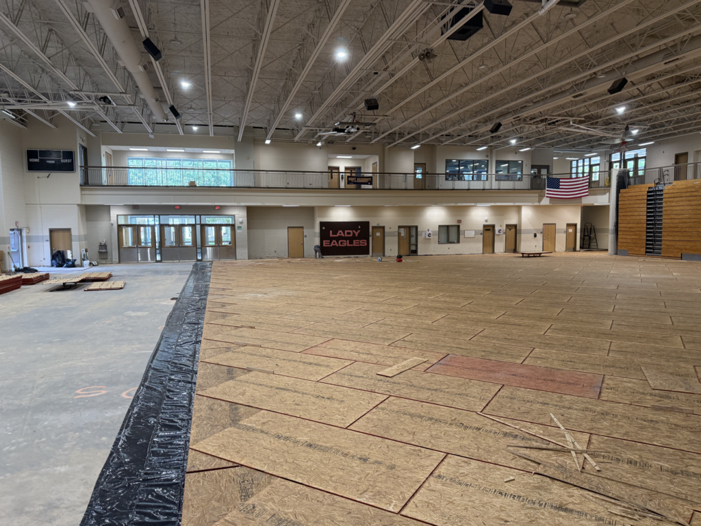 Indoor gymnasium with plywood panels covering the floor during renovation, bleachers on the right, and an American flag hanging near the ceiling.