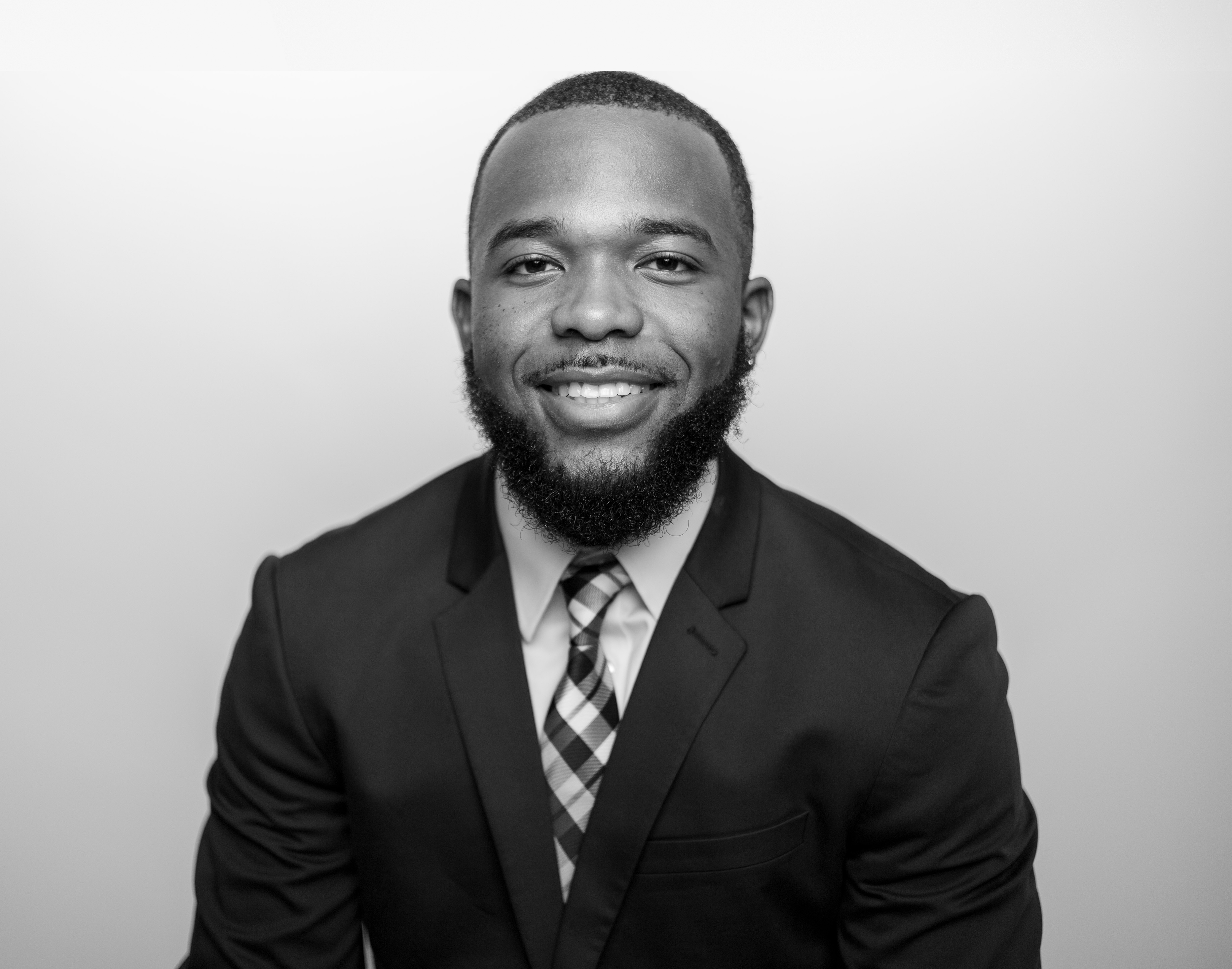 Smiling man with a beard wearing a suit jacket and plaid tie against a plain background.